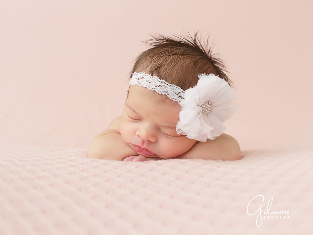 Newborn baby girl wearing a white flower headband and laying on a pink blanket