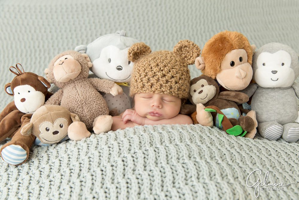 newborn baby boy wearing a knitted teddy bear beanie and laying with his stuffed animal toys