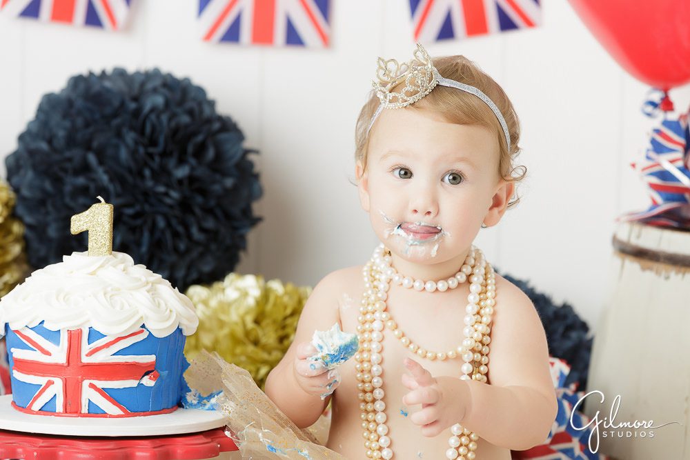 Princess London's 1st Birthday, eating, cake smash, smiling, portraits, photography, family, england theme, outfit, skirt, tutu, headband, necklaces, one year old, pom poms, flags, props