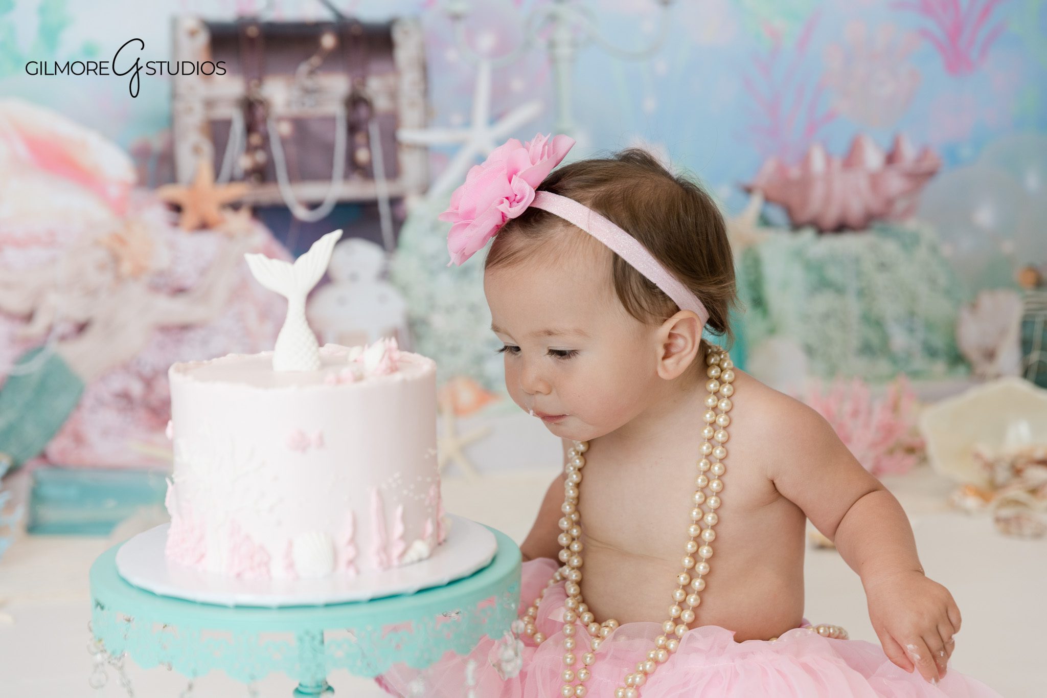 Baby girl smiling beside pink birthday cake on aqua cake stand
Newport Beach baby cake smash session with pearl and mermaid theme