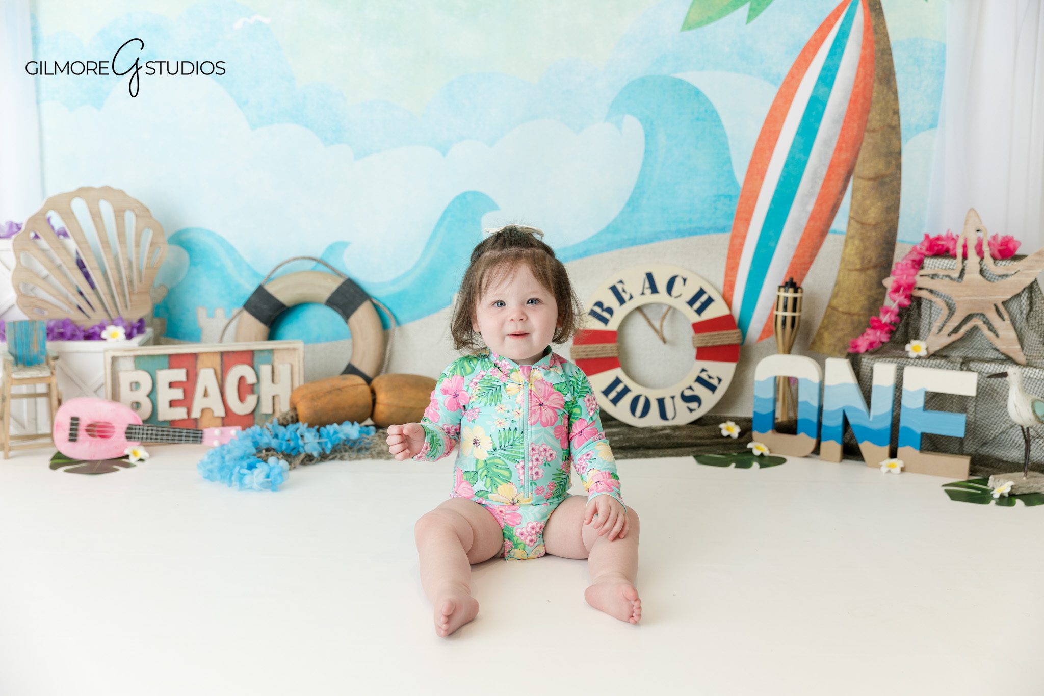 Beach house first birthday setup with white cake and plumeria flowers
Baby girl sitting in beach theme birthday set with one sign