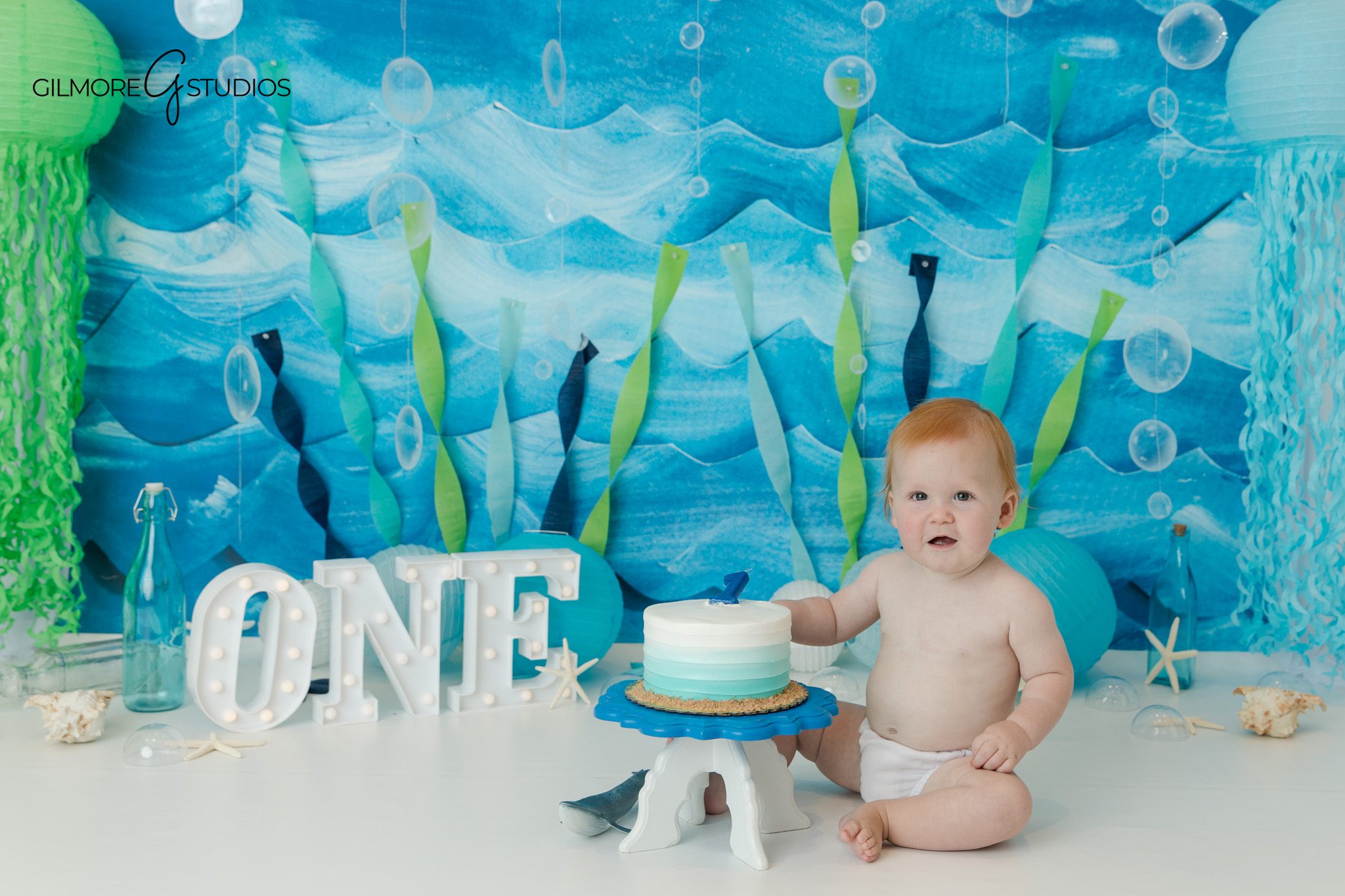 First birthday boy with red hair smiling during Queen Creek studio photos
Baby boy sitting beside blue and silver first birthday balloons